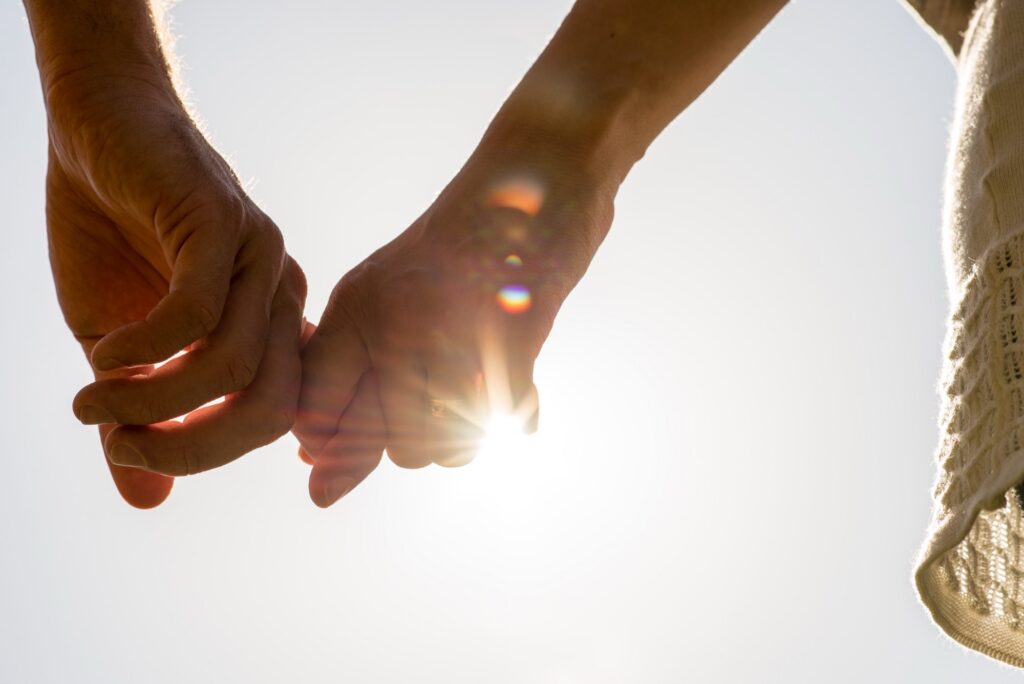 Close up Hands of Romantic Couple Holding Together with Sun Rays on a White Sky.
